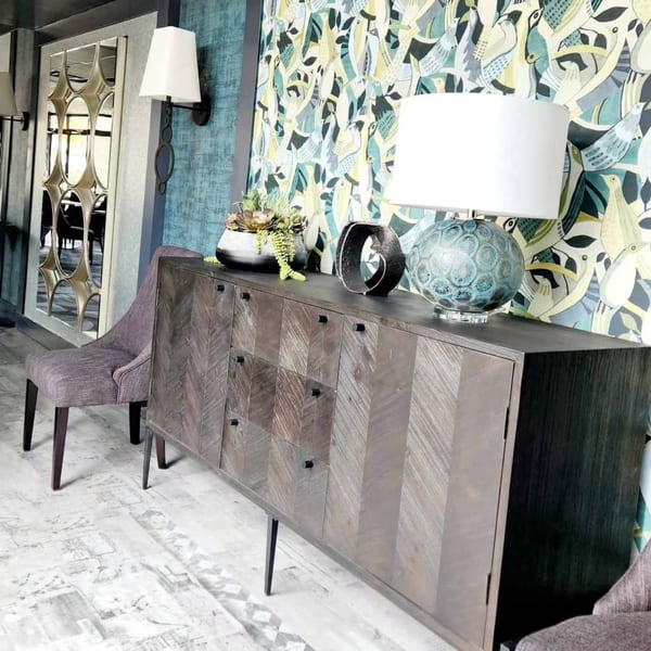 A modern wooden cabinet with a lamp and plant sits against a patterned wallpaper wall.